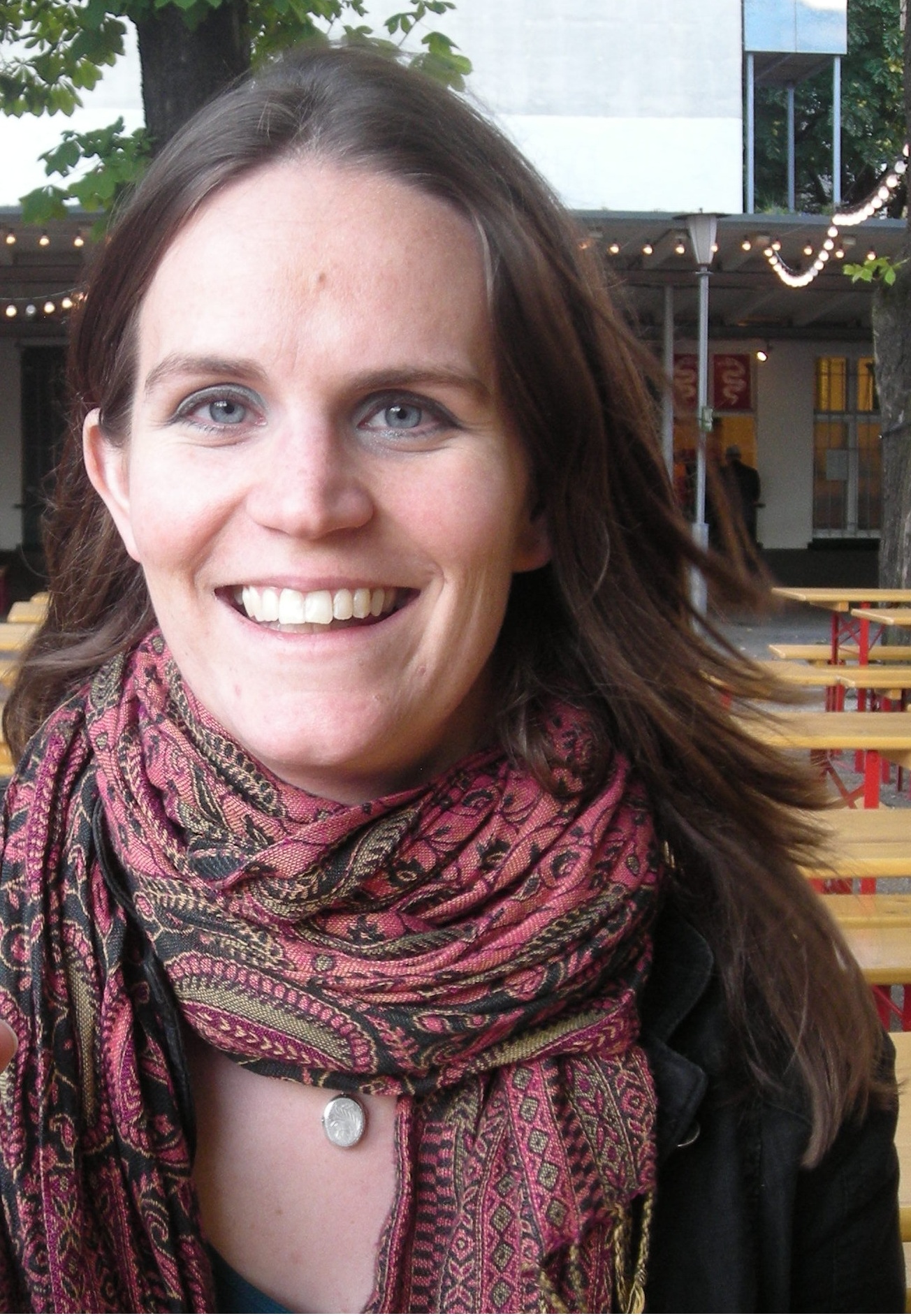 Picture of Helen. She is a white woman with long brown hair, wearing a scarf and jacket and sitting outdoors. There are tables and strings of lights behind her. 